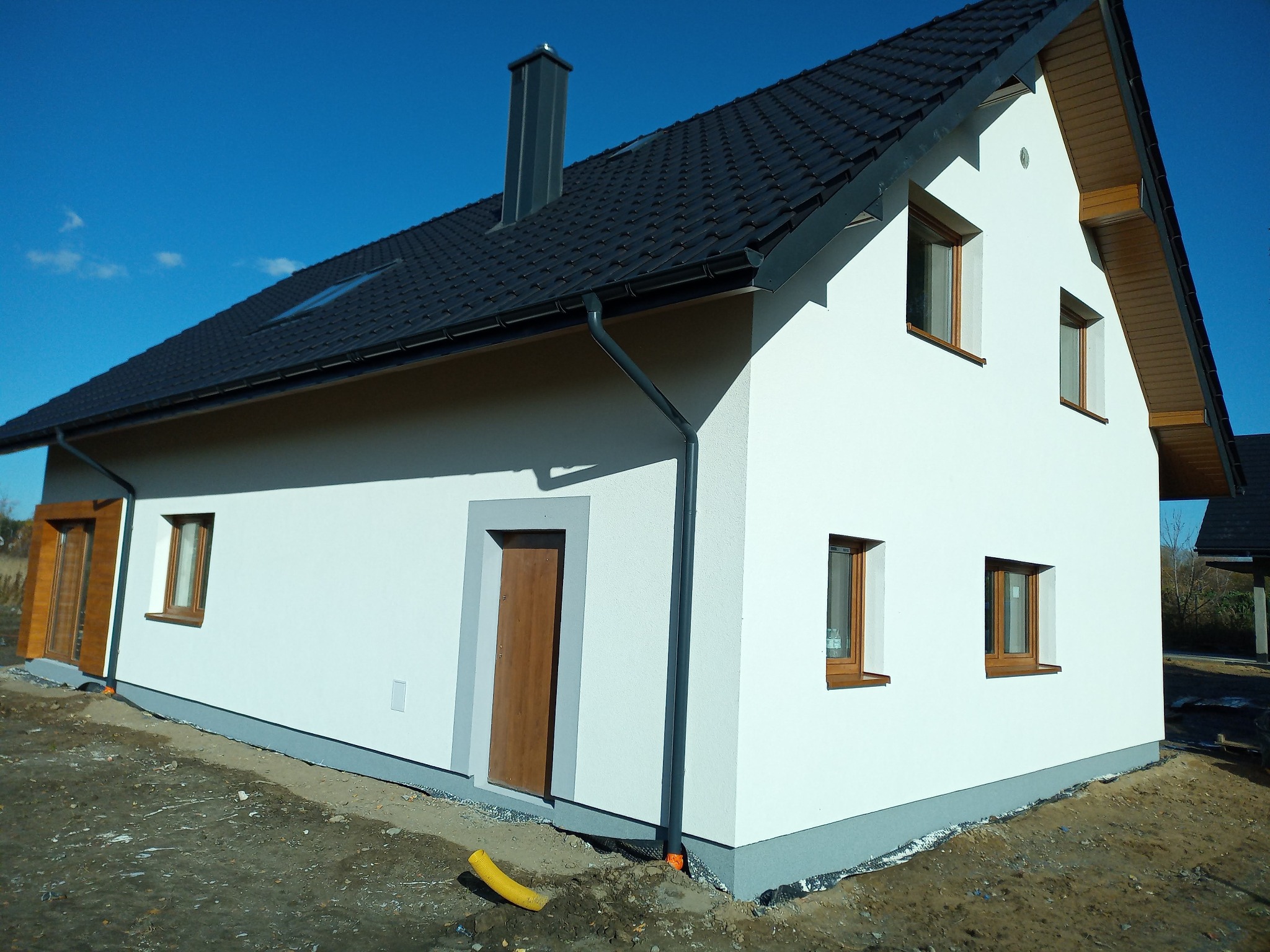 Nowoczesny dom jednorodzinny z białą elewacją i ciemną dachówką, widok z boku na świeżo wykończoną ścianę z brązowymi oknami i drzwiami, żółta rura drenażowa leżąca obok fundamentu.