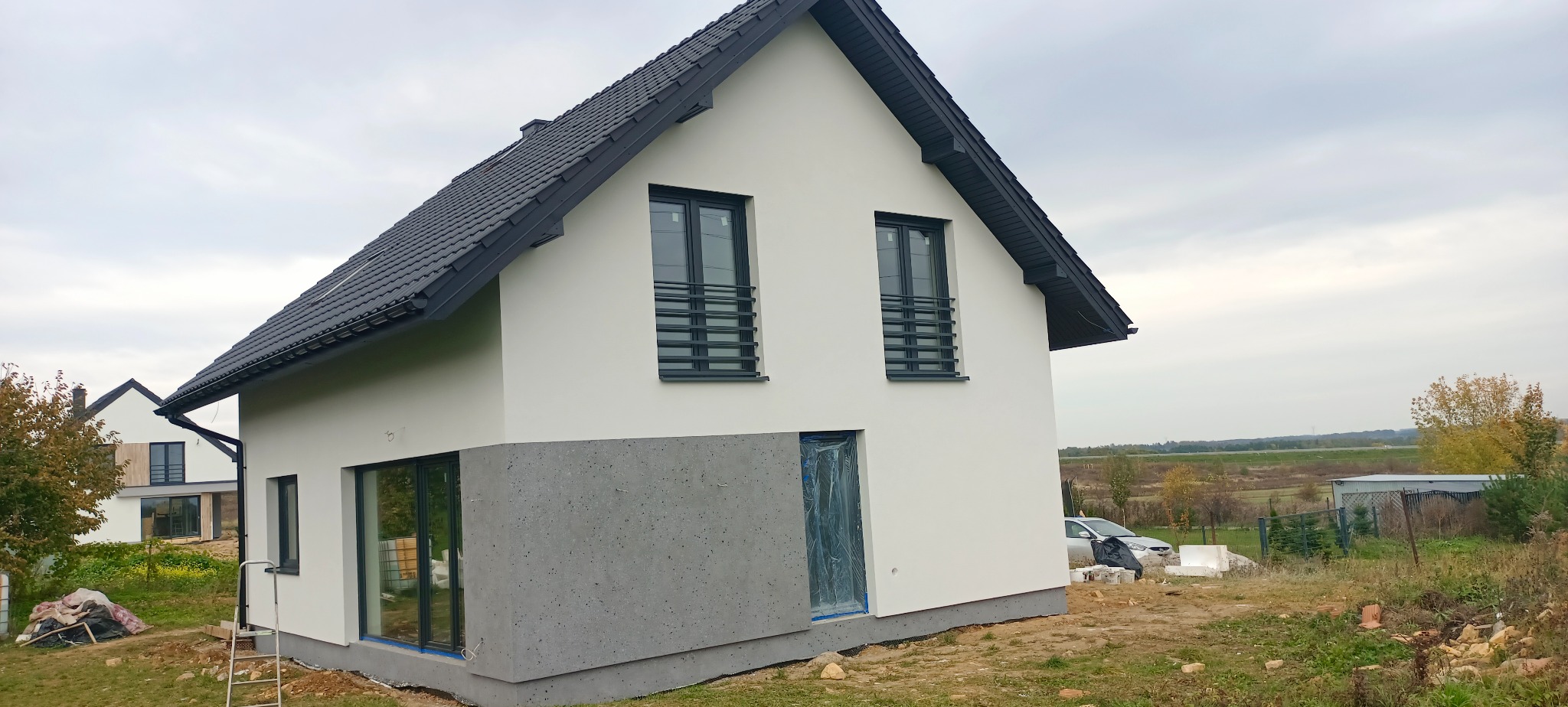 Nowoczesny dom jednorodzinny z białą elewacją i szarym cokołem, okna zabezpieczone folią, widoczny fragment ogrodu i krajobraz w tle.