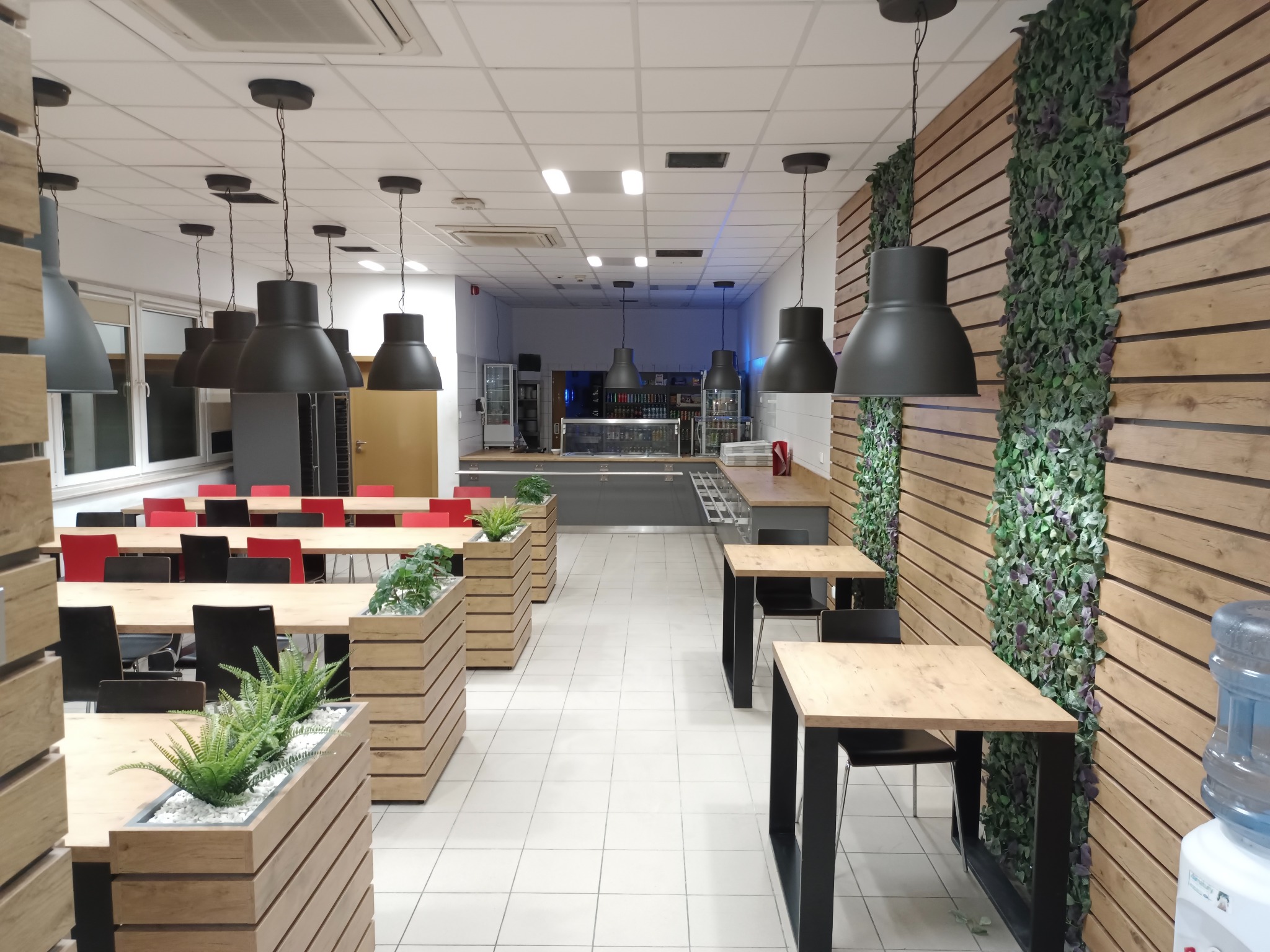 Nowoczesna aranżacja wnętrza biurowej stołówki z drewnianymi panelami ściennymi, roślinami w donicach i wiszącymi lampami w stylu industrialnym.