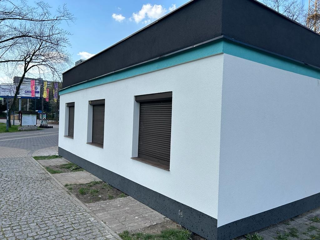 Nowo wykończony budynek z białą elewacją, trzema zamkniętymi oknami z brązowymi roletami, ciemnoszarym dachem i podmurówką, widoczny chodnik z kostki brukowej.