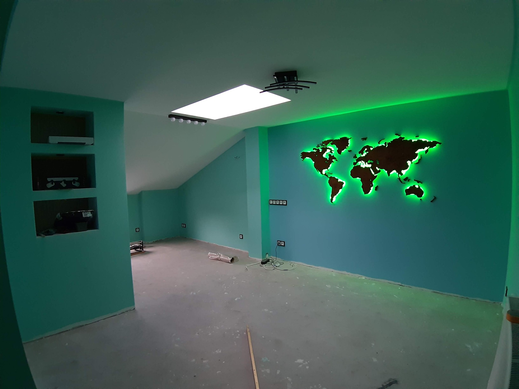 Turkusowe ściany w pokoju na poddaszu z wnękami, podświetlana mapa świata na ścianie, biały sufit z oknem dachowym i nowoczesnym oświetleniem, szara wylewka na podłodze w trakcie prac wykończeniowych.