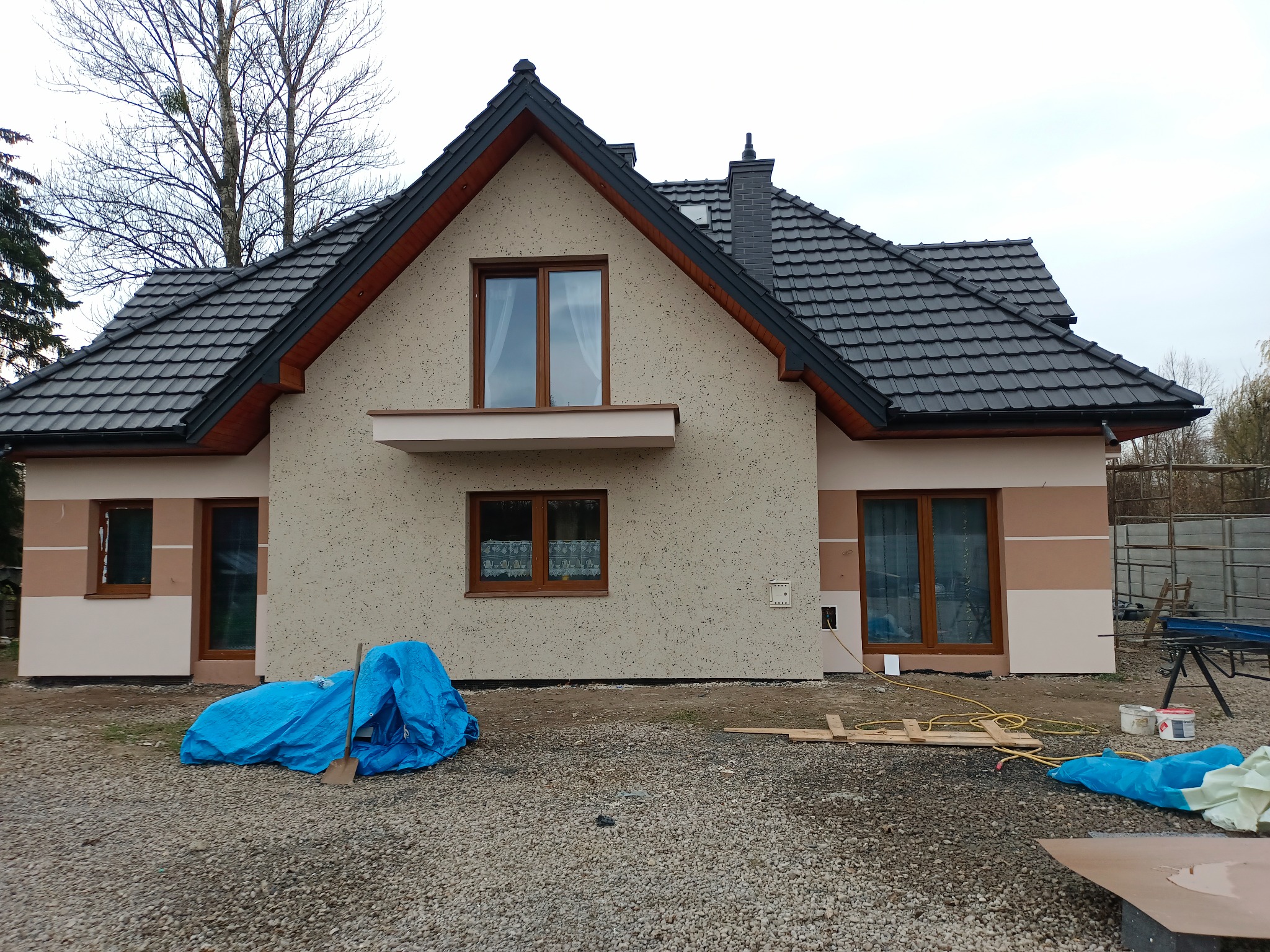 Dwukondygnacyjny dom jednorodzinny z szarą dachówką i elewacją w odcieniach beżu, z widocznymi śladami prac wykończeniowych i narzędziami budowlanymi na posesji.