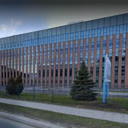 Nowoczesny budynek biurowy z ceglaną elewacją i szklanymi oknami, widok z zewnątrz, z chodnikiem i zielenią w tle.