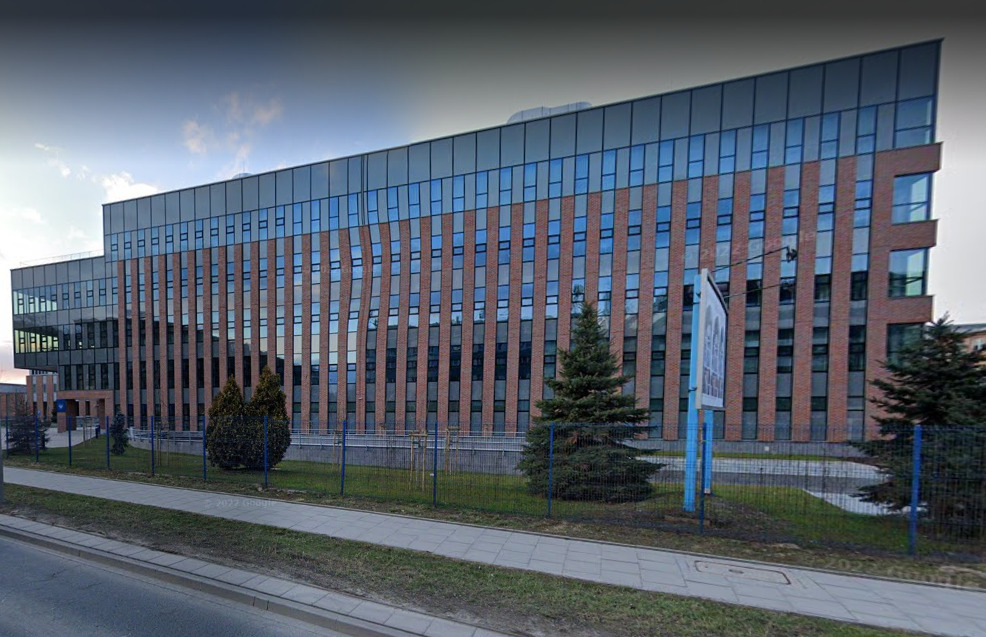 Nowoczesny budynek biurowy z ceglaną elewacją i szklanymi oknami, widok z zewnątrz, z chodnikiem i zielenią w tle.