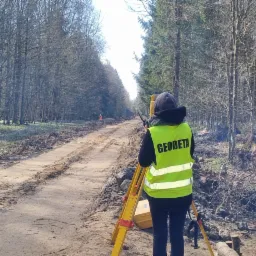 Osoba w odblaskowej kamizelce z napisem 'GEODETA' obsługuje niwelator na tle leśnej drogi w trakcie prac pomiarowych.