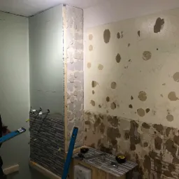 Łazienka w trakcie układania szarej mozaiki na ścianie, widoczne poziomice i przygotowane ściany do dalszych prac wykończeniowych.