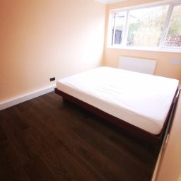 Meb-art - Pomalowane na brzoskwiniowo ściany pokoju z ciemną podłogą z paneli, białym sufitem, grzejnikiem pod oknem i łóżkiem z białym materacem na drewnianej ramie.