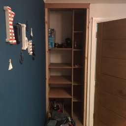 Meb-art - Wnęka w ścianie przygotowana pod zabudowę szafy w pokoju dziecięcym, widoczne półki z surowego drewna, zabawki na półkach, dekoracje ścienne w stylu marynistycznym, kosz z ubraniami u dołu.