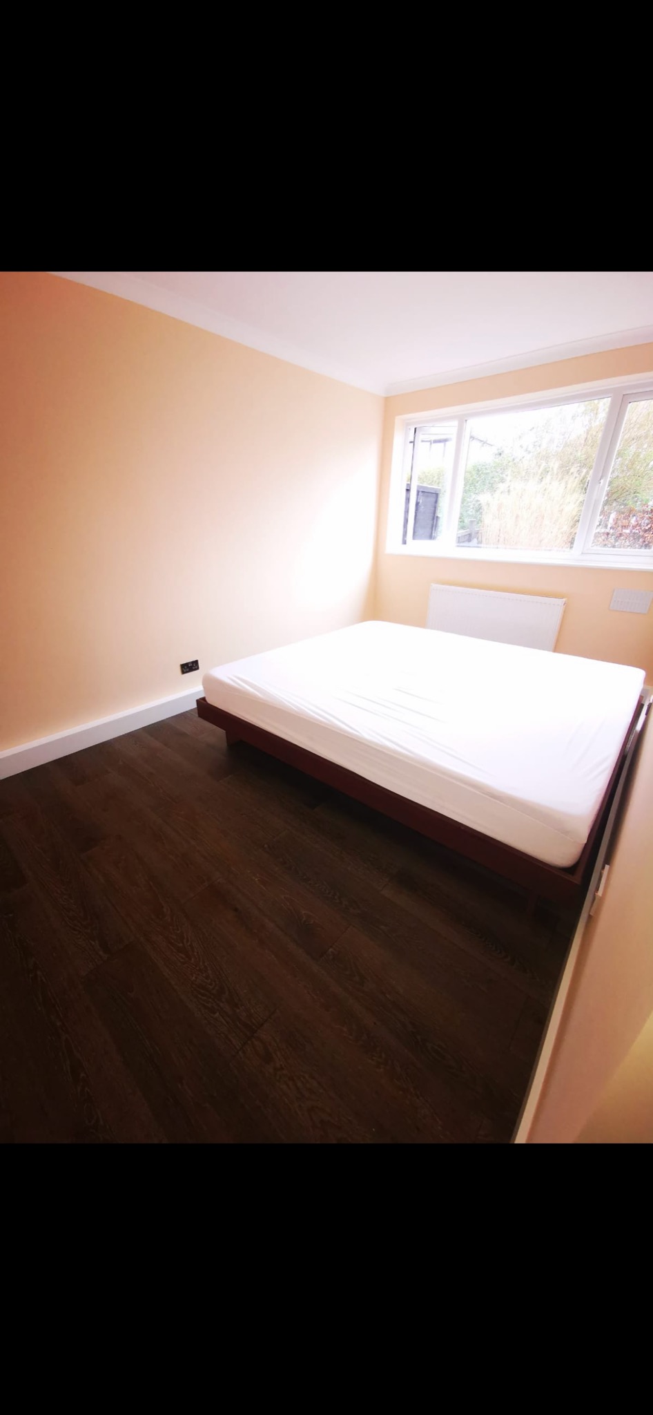 Pomalowane na brzoskwiniowo ściany pokoju z ciemną podłogą z paneli, białym sufitem, grzejnikiem pod oknem i łóżkiem z białym materacem na drewnianej ramie.