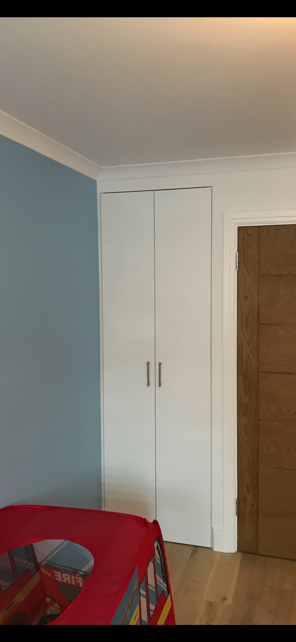 Wbudowana biała szafa z dwoma drzwiami i metalowymi uchwytami, na tle błękitnej ściany i drewnianej podłogi z fragmentem czerwonego namiotu dla dzieci w kształcie wozu strażackiego w rogu