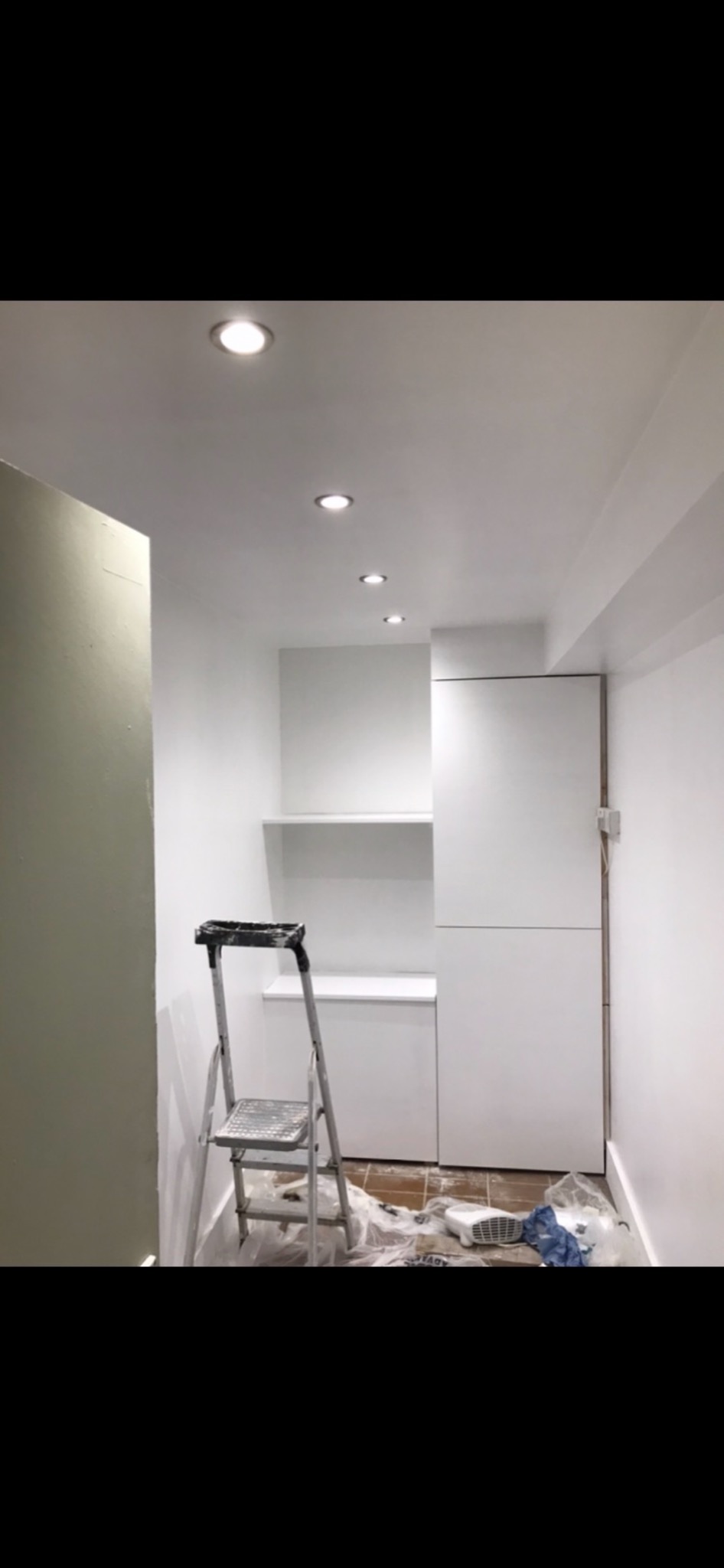 Wnętrze w trakcie prac wykończeniowych: pomalowane na biało ściany, zabudowa meblowa z półkami, drabina malarska i folia ochronna na podłodze.