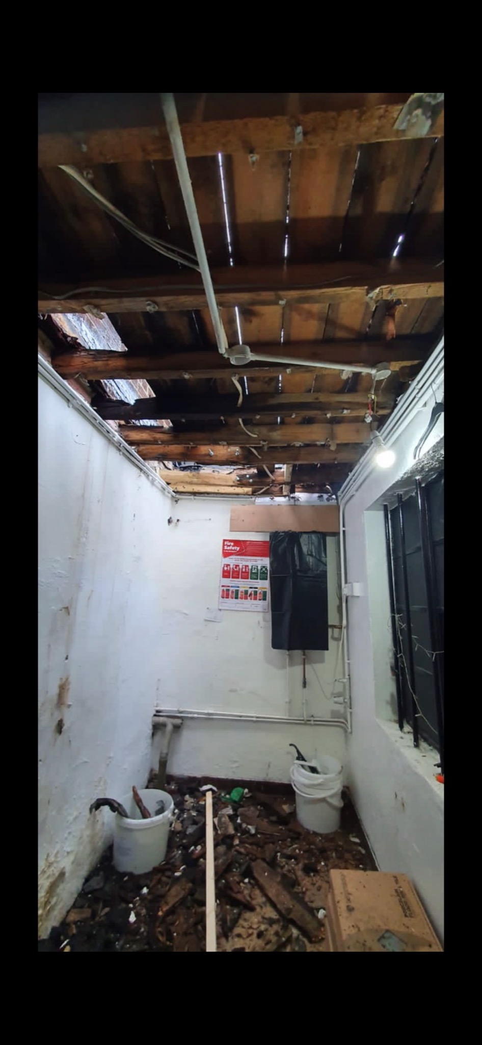 Piwnica w trakcie demontażu sufitu, widoczne odsłonięte belki stropowe, instalacje elektryczne, gruz na podłodze, białe ściany z widocznymi śladami wilgoci, plakat informacyjny na ścianie.