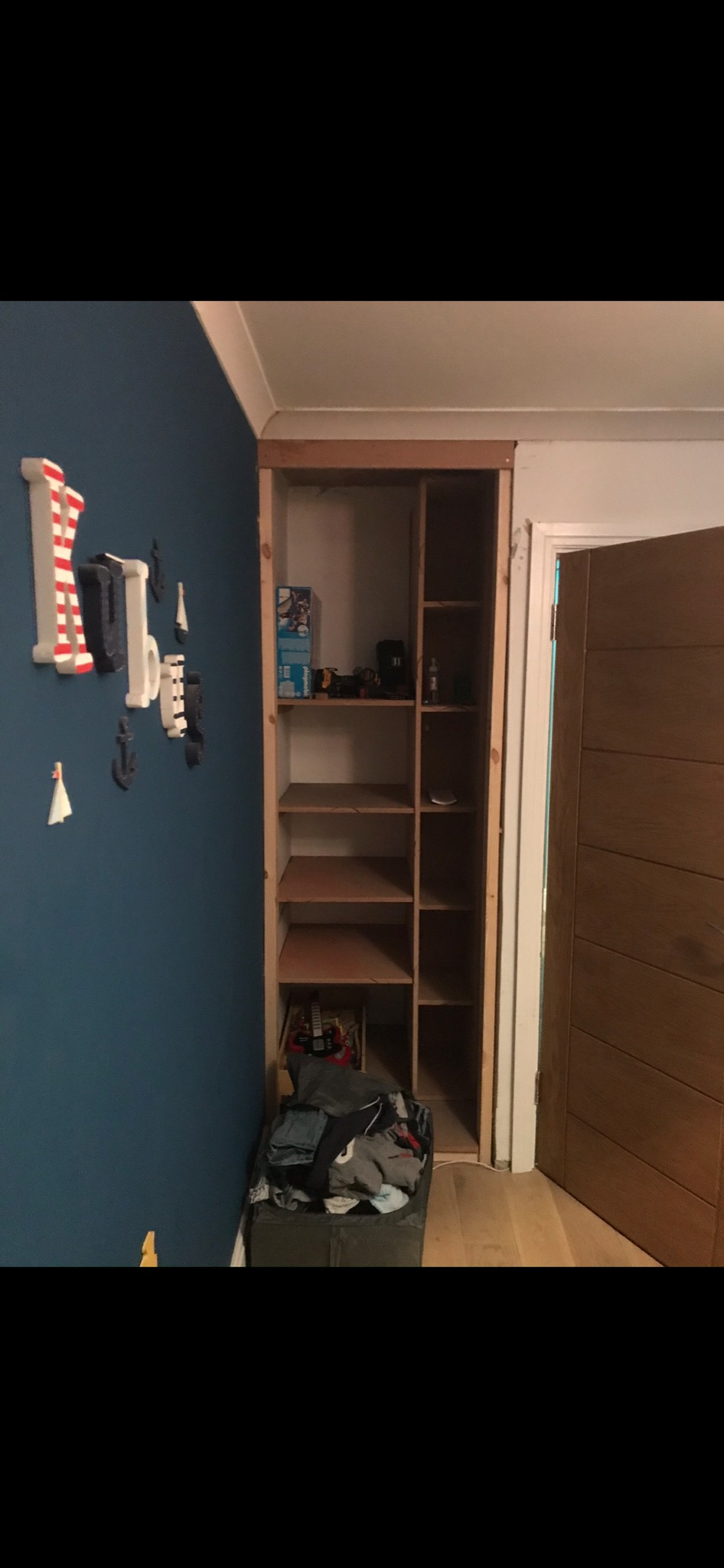 Wnęka w ścianie przygotowana pod zabudowę szafy w pokoju dziecięcym, widoczne półki z surowego drewna, zabawki na półkach, dekoracje ścienne w stylu marynistycznym, kosz z ubraniami u dołu.