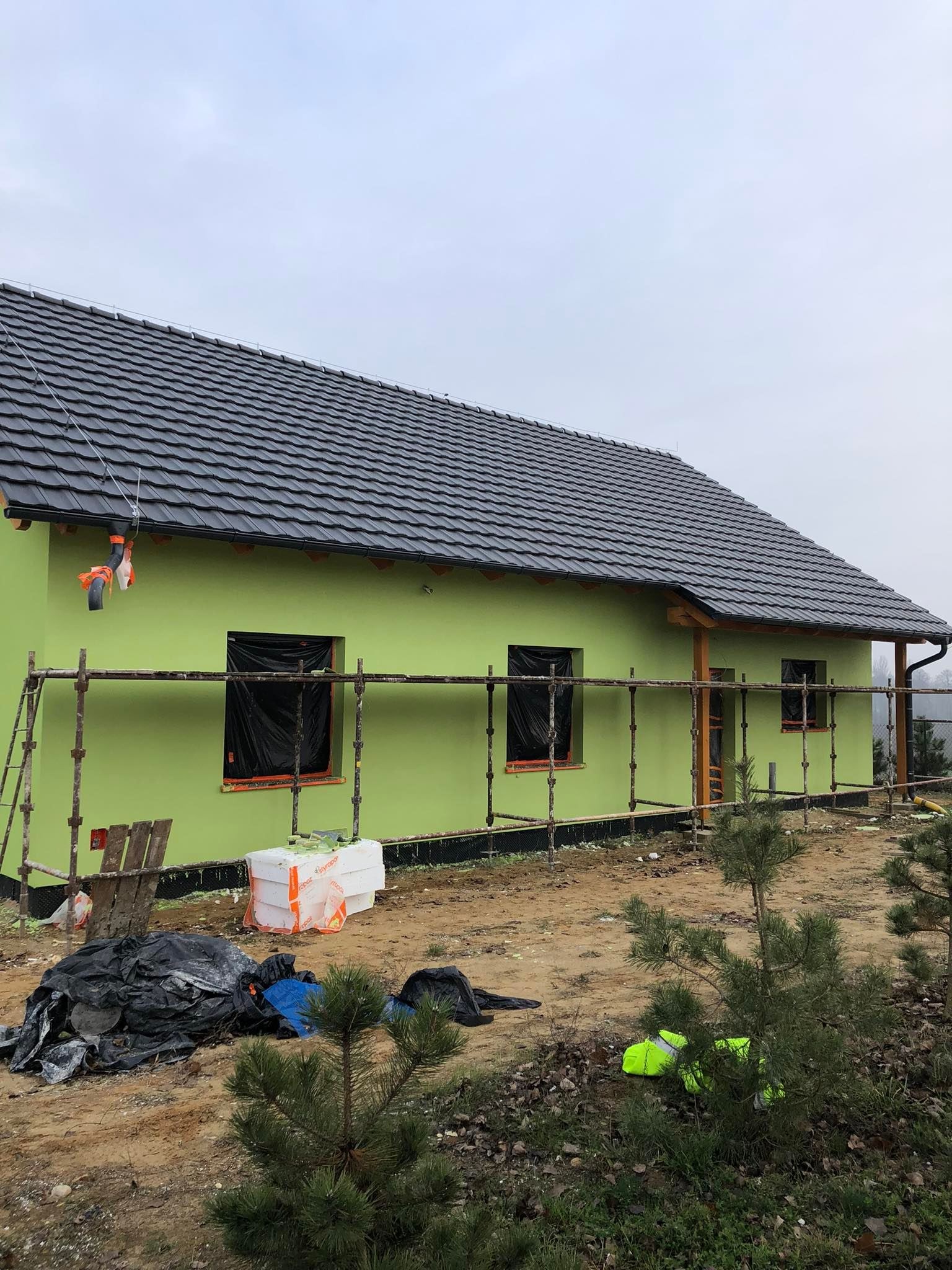 Dom jednorodzinny w trakcie prac elewacyjnych, z zieloną, świeżo nałożoną warstwą tynku i rusztowaniami. Okna zabezpieczone folią, widoczne materiały budowlane i teren wokół domu.