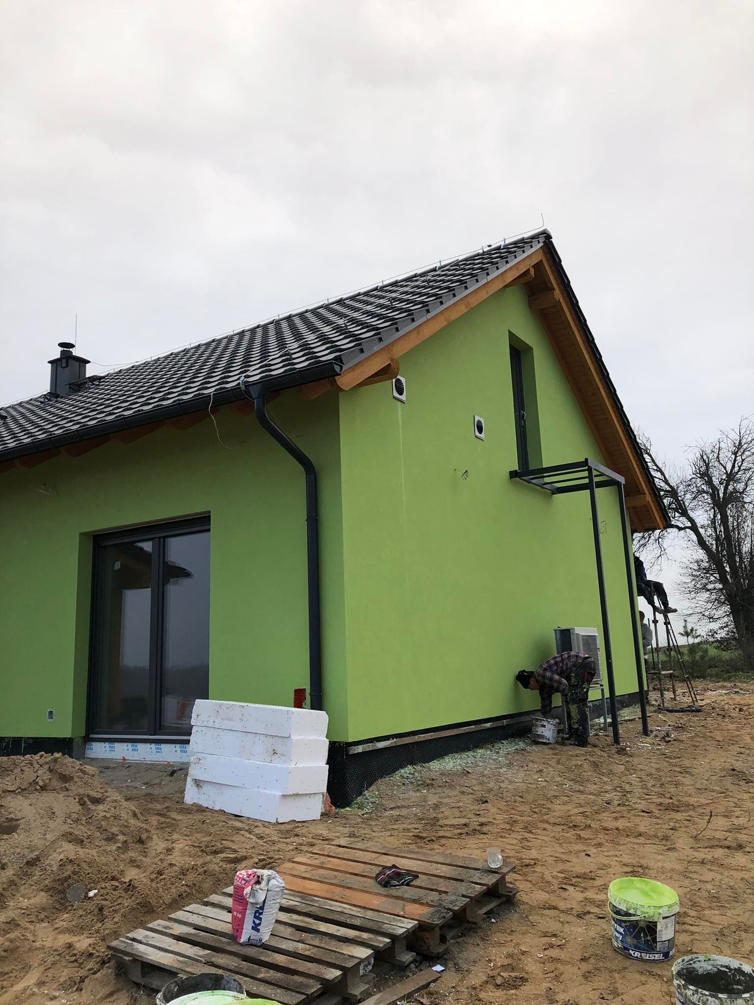 Świeżo otynkowany, jasnozielony dom jednorodzinny z czarną dachówką, widoczny pracownik na drabinie nanoszący poprawki, styropian i wiaderka z farbą leżące na ziemi.