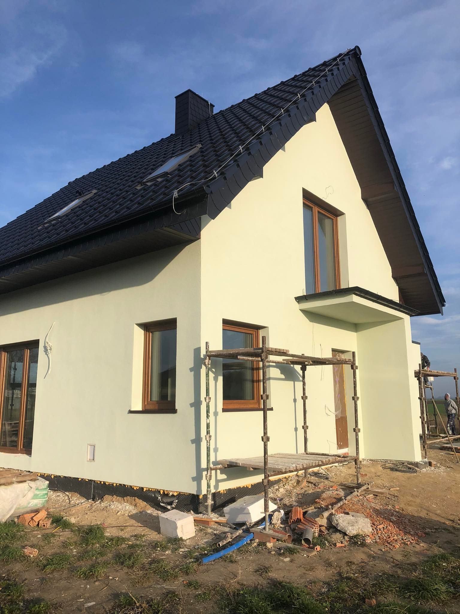 Dom jednorodzinny po nałożeniu jasnej elewacji, widoczne rusztowania i teren wokół domu w trakcie prac wykończeniowych, czarny dach z dachówki.