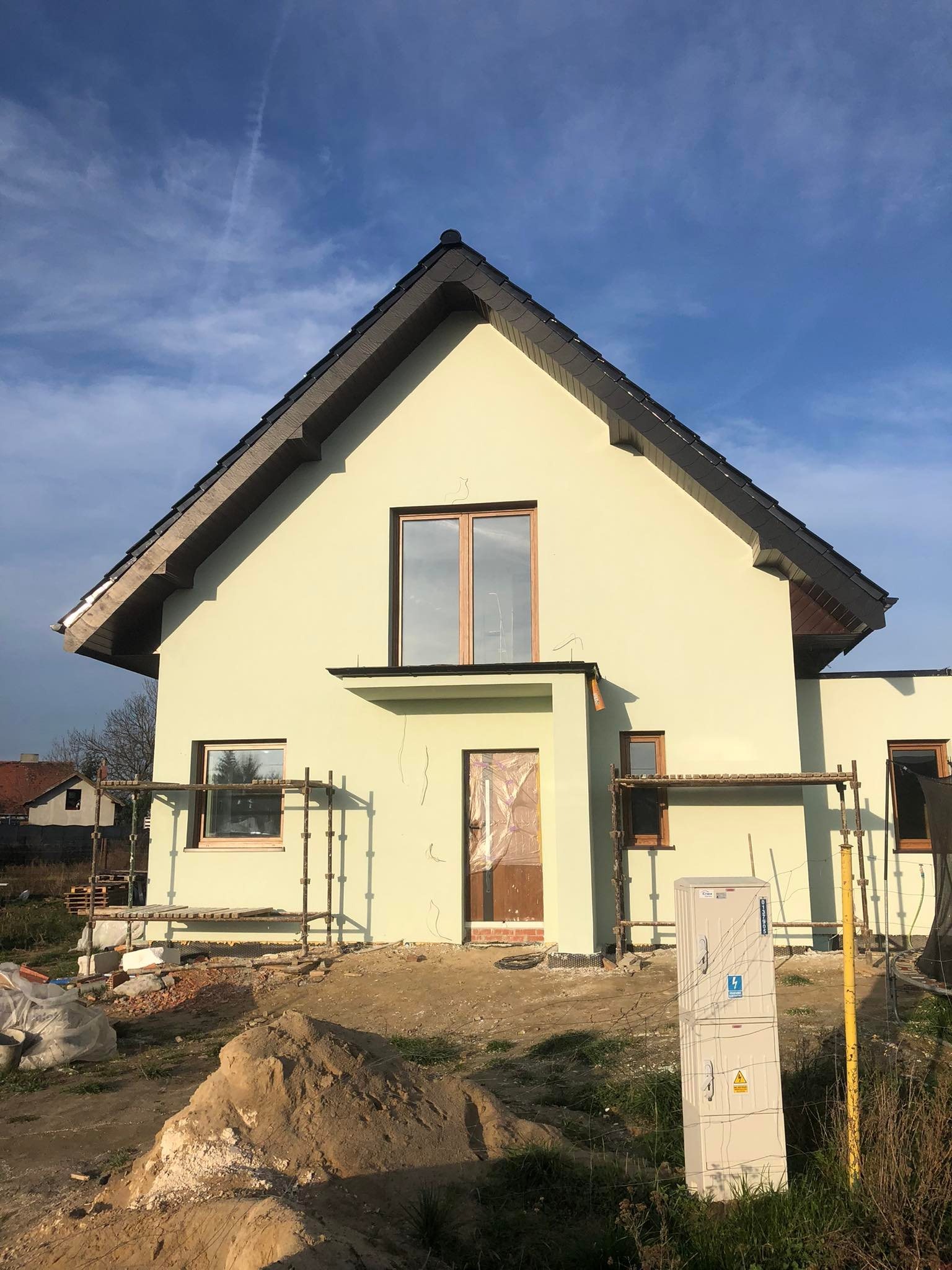 Dom jednorodzinny z jasną elewacją w trakcie wykańczania; widoczne rusztowania, okna z drewnianą stolarką, niezabezpieczone wejście, skrzynka elektryczna i sterta piachu na pierwszym planie.