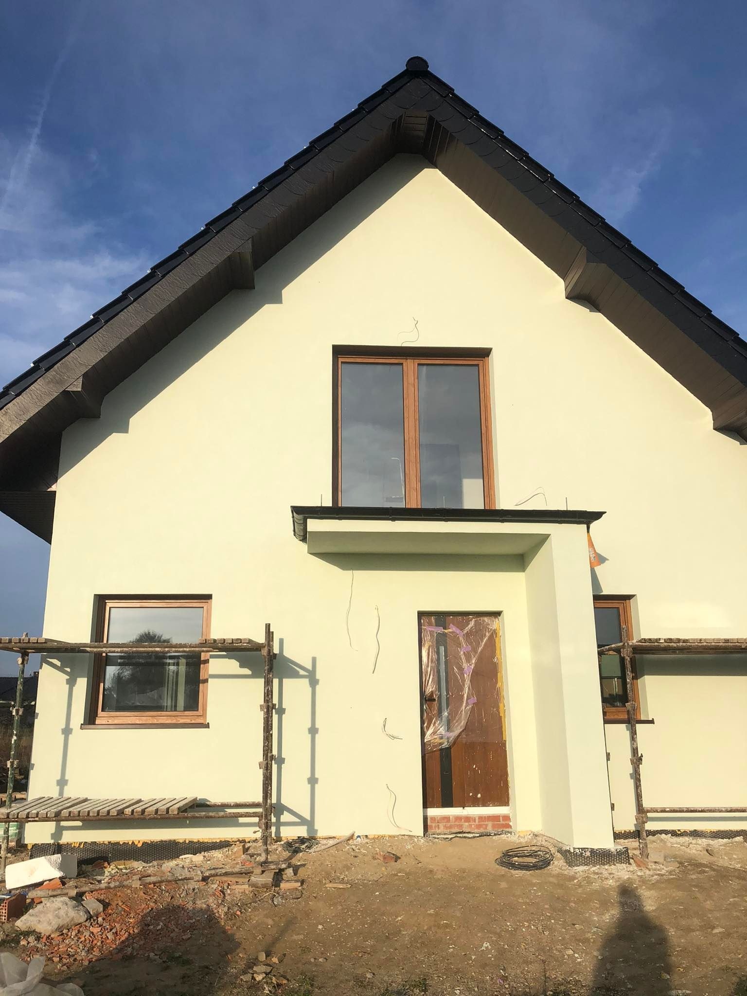 Dom jednorodzinny z jasną elewacją w trakcie prac wykończeniowych, widoczne rusztowania i zabezpieczone folią drzwi wejściowe, świeżo otynkowana ściana zewnętrzna z widocznymi pęknięciami.