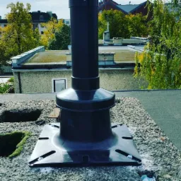 Czarny komin wentylacyjny zamontowany na płaskim dachu pokrytym żwirem, widoczne śruby mocujące i wycięte otwory w podłożu, w tle budynki mieszkalne i drzewa.