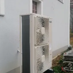 Zewnętrzna jednostka pompy ciepła Hitachi zamontowana na betonowych bloczkach fundamentowych przy białej ścianie budynku z oknami.