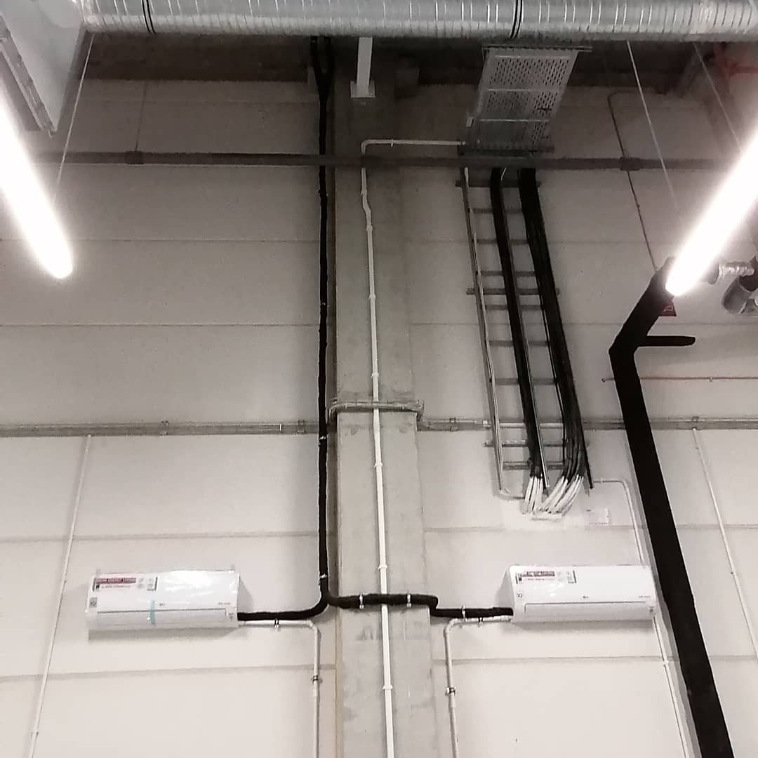 Dwa białe klimatyzatory LG zamontowane na ścianie hali przemysłowej, widoczne rury i kable poprowadzone wzdłuż betonowego słupa i pod sufitem, drabina serwisowa w tle.