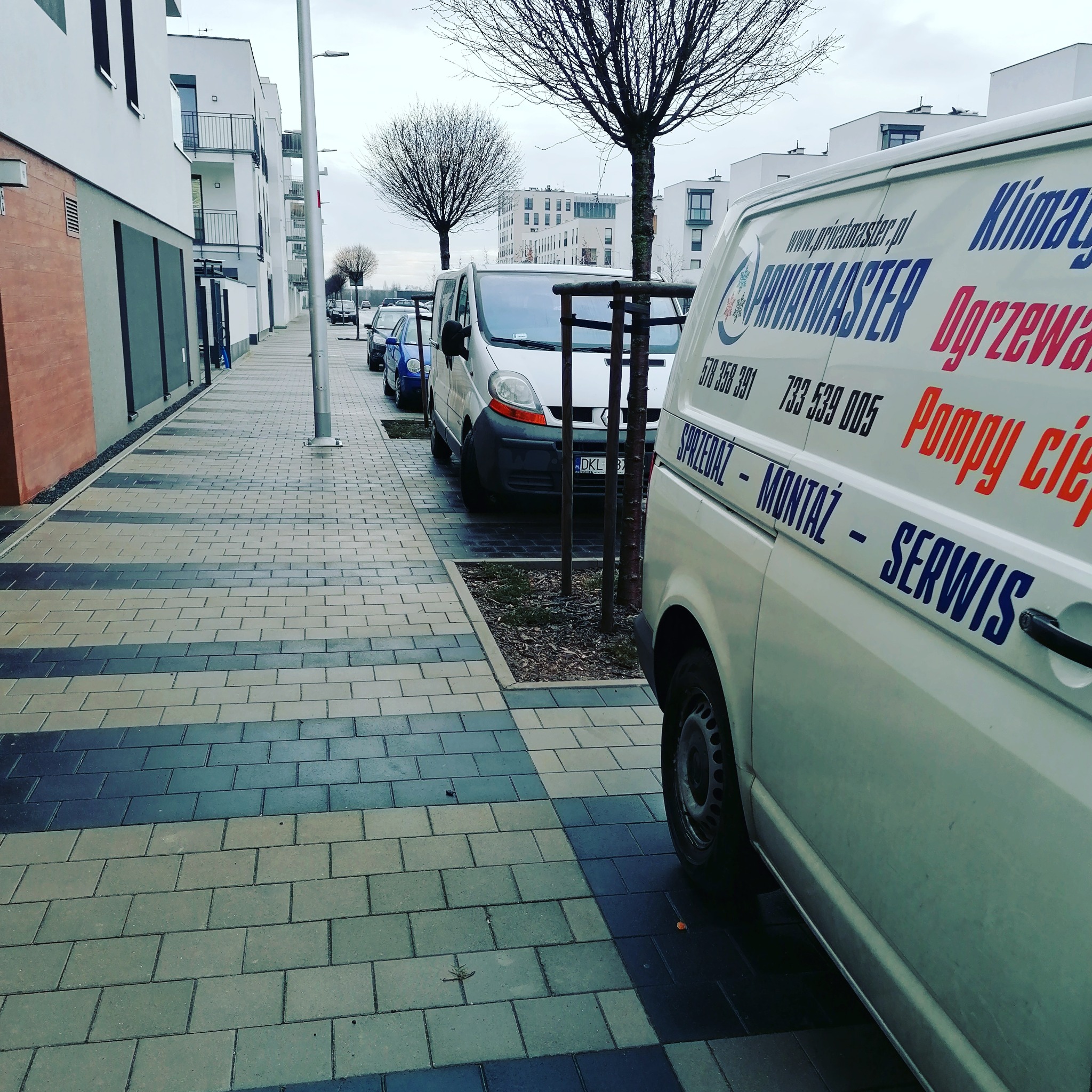 Samochód dostawczy z reklamą firmy świadczącej usługi montażu i serwisu klimatyzacji, ogrzewania i pomp ciepła, zaparkowany na chodniku przed nowoczesnym budynkiem mieszkalnym.