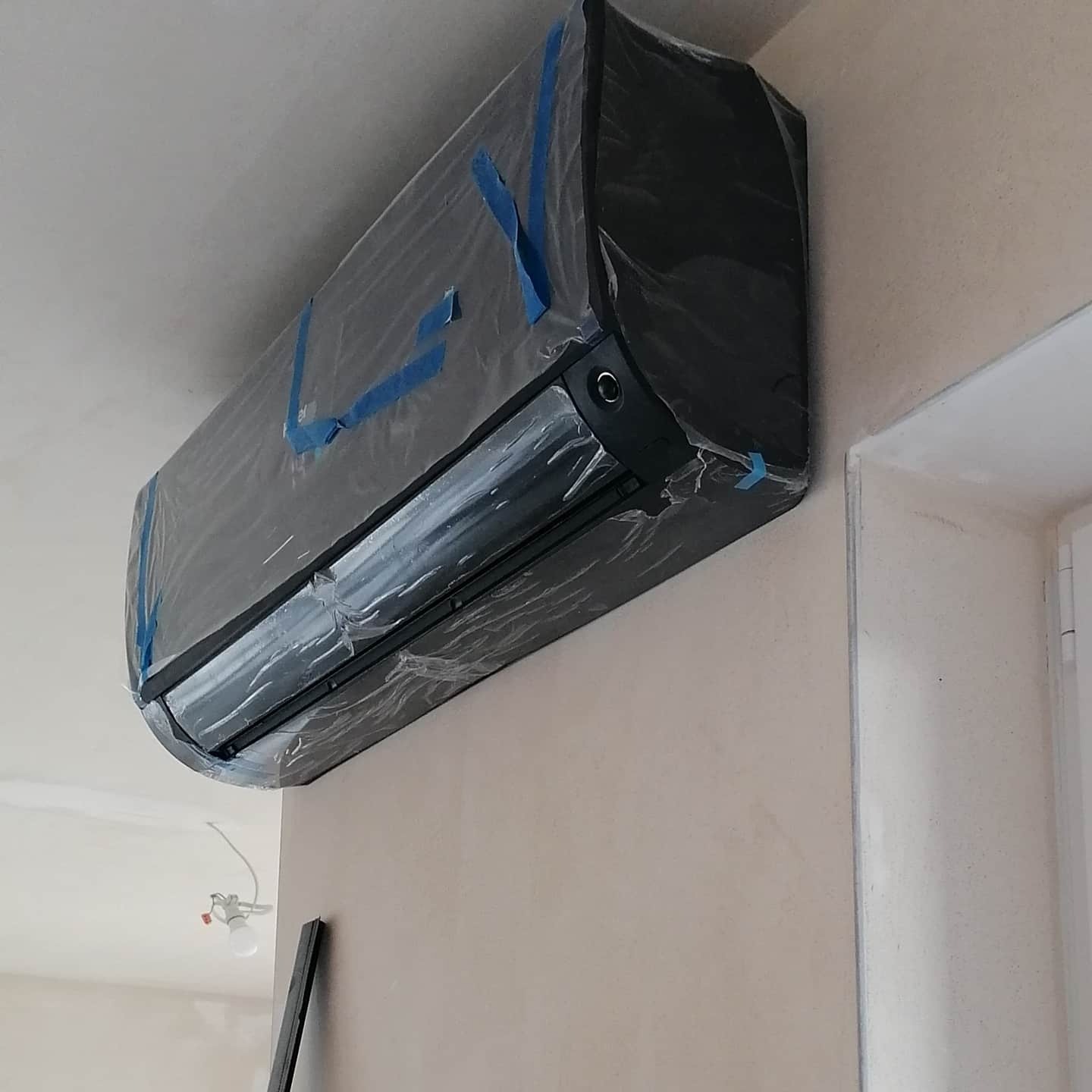 Czarny klimatyzator ścienny zabezpieczony folią ochronną i niebieską taśmą malarską, zamontowany na beżowej ścianie w trakcie remontu, widoczna surowa żarówka na suficie.