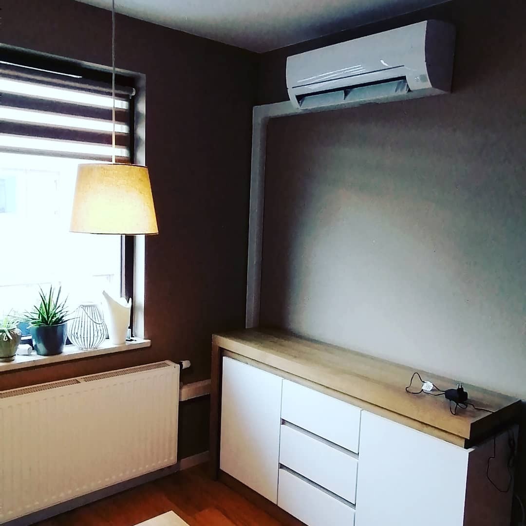 Biała klimatyzacja ścienna zamontowana w nowoczesnym salonie z brązowymi ścianami, widoczny biały grzejnik pod oknem z roletami i drewniana komoda z białymi frontami.