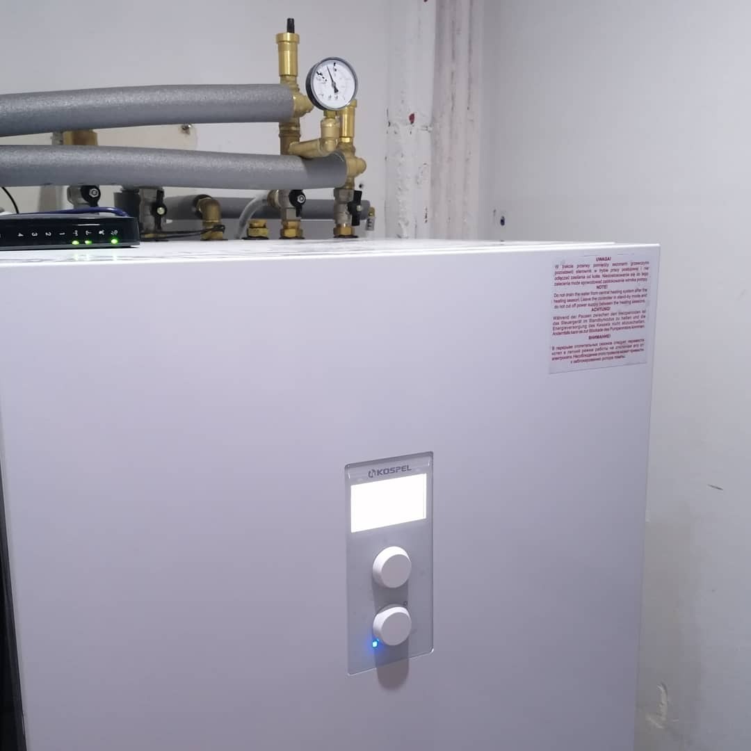 Biały piec gazowy Kospel z widocznym manometrem, rurami w izolacji i panelem sterowania z dwoma pokrętłami, umieszczony na tle białej ściany z widocznymi śladami użytkowania.