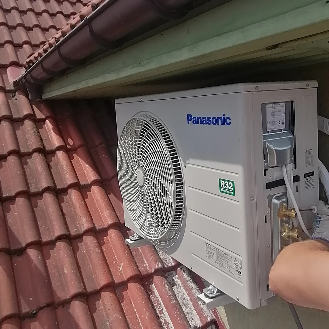 Zewnętrzna jednostka klimatyzacji Panasonic R32 zamontowana na dachu z czerwonej dachówki, widoczne przyłącza i ręka instalatora.