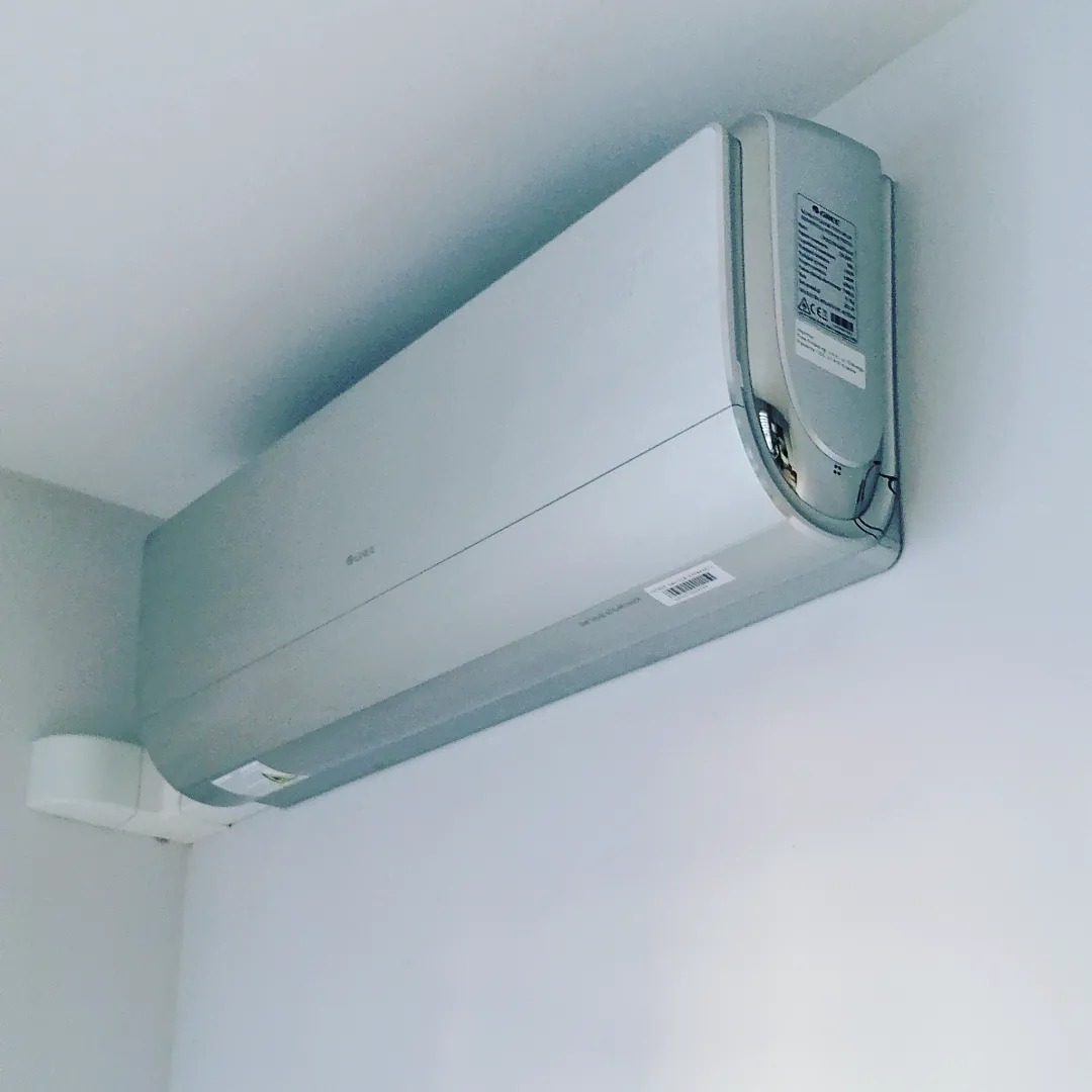 Zamontowany, srebrny klimatyzator marki Gree na białej ścianie w rogu pomieszczenia, widoczna biała maskownica instalacji elektrycznej.