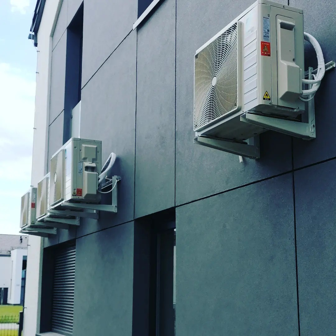 Zewnętrzne jednostki klimatyzacji zamontowane na elewacji budynku z szarego panelu kompozytowego, widoczne okna i rolety, ujęcie z dołu.