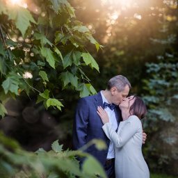 FOTSWIM MICHAŁ DEREWECKI - Para całująca się w plenerze, mężczyzna w granatowym garniturze z muchą, kobieta w białej sukience. Ciepłe światło słoneczne, zielone liście w tle.