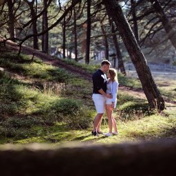 FOTSWIM MICHAŁ DEREWECKI - Zakochana para w lesie, mężczyzna całuje kobietę w czoło. Ujęcie zza pnia drzewa, miękkie światło słoneczne, naturalna sceneria. Romantyczna atmosfera.