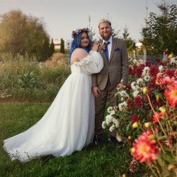 FOTSWIM MICHAŁ DEREWECKI - Para młoda w plenerze: panna młoda w białej sukni z niebieskimi włosami i wiankiem, pan młody w brązowym garniturze. Kwiaty w tle.