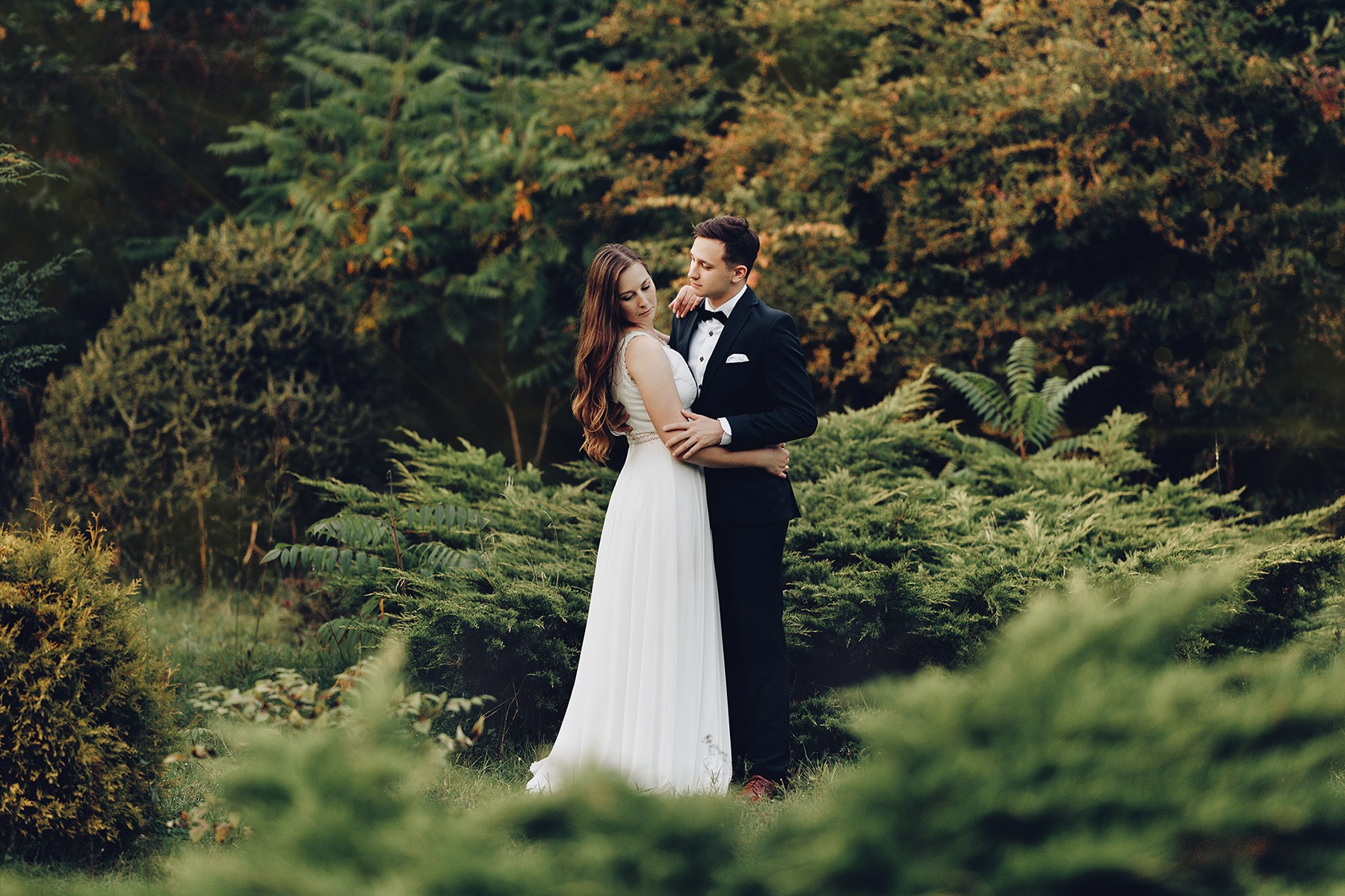 Elegancka para młoda obejmująca się w malowniczym ogrodzie pełnym zieleni, on w klasycznym smokingu z muchą, ona w zwiewnej białej sukni z koronkowym pasem, oboje zamyśleni.