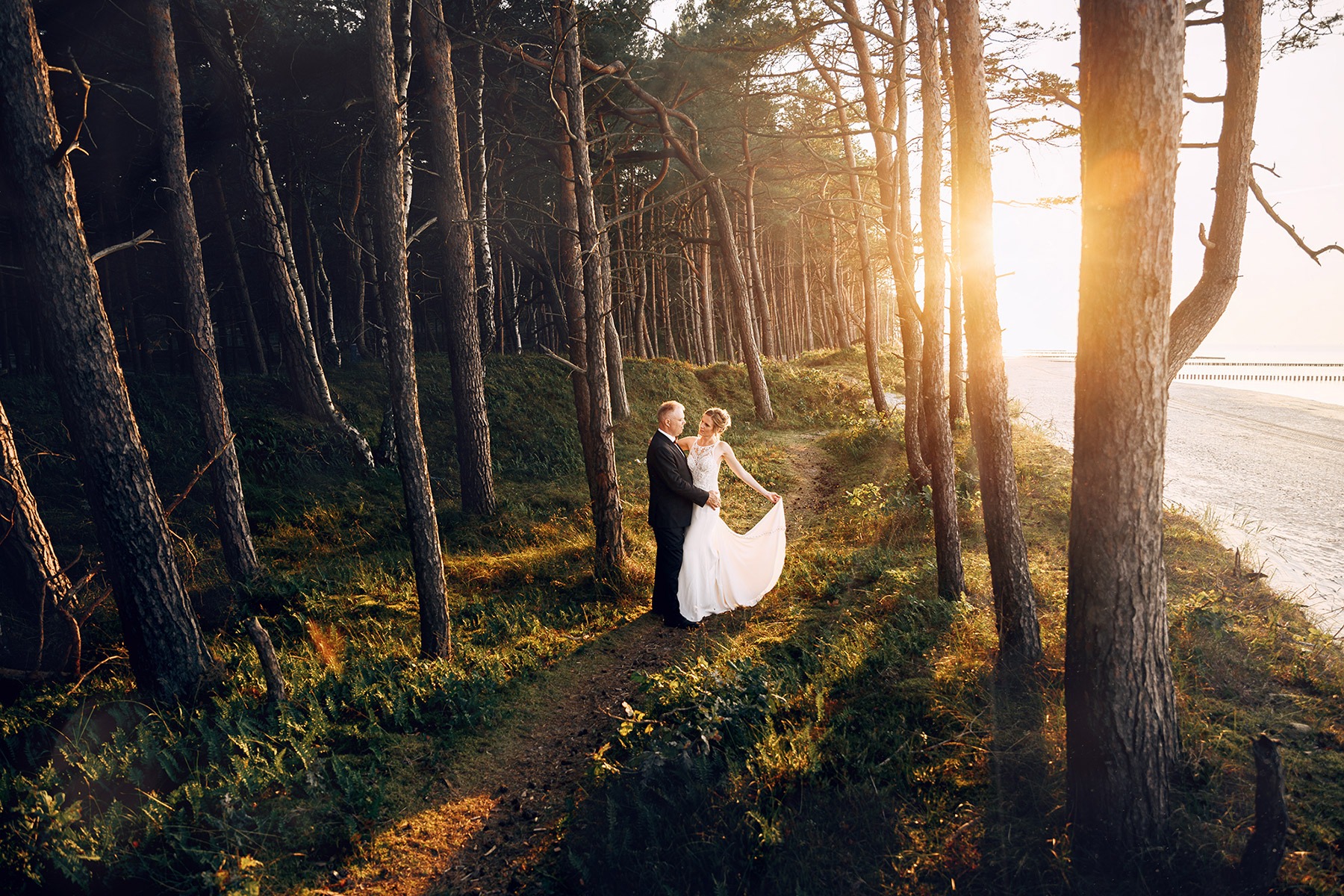 Para młoda w lesie sosnowym w promieniach zachodzącego słońca, panna młoda w sukni z koronkową górą trzyma brzeg sukni, w tle plaża i morze.