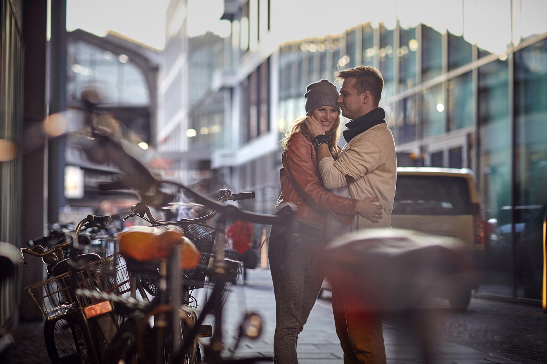 Uśmiechnięta kobieta w czapce i skórzanej kurtce, obejmowana przez mężczyznę na tle miejskiej ulicy z rowerami i nowoczesną architekturą.