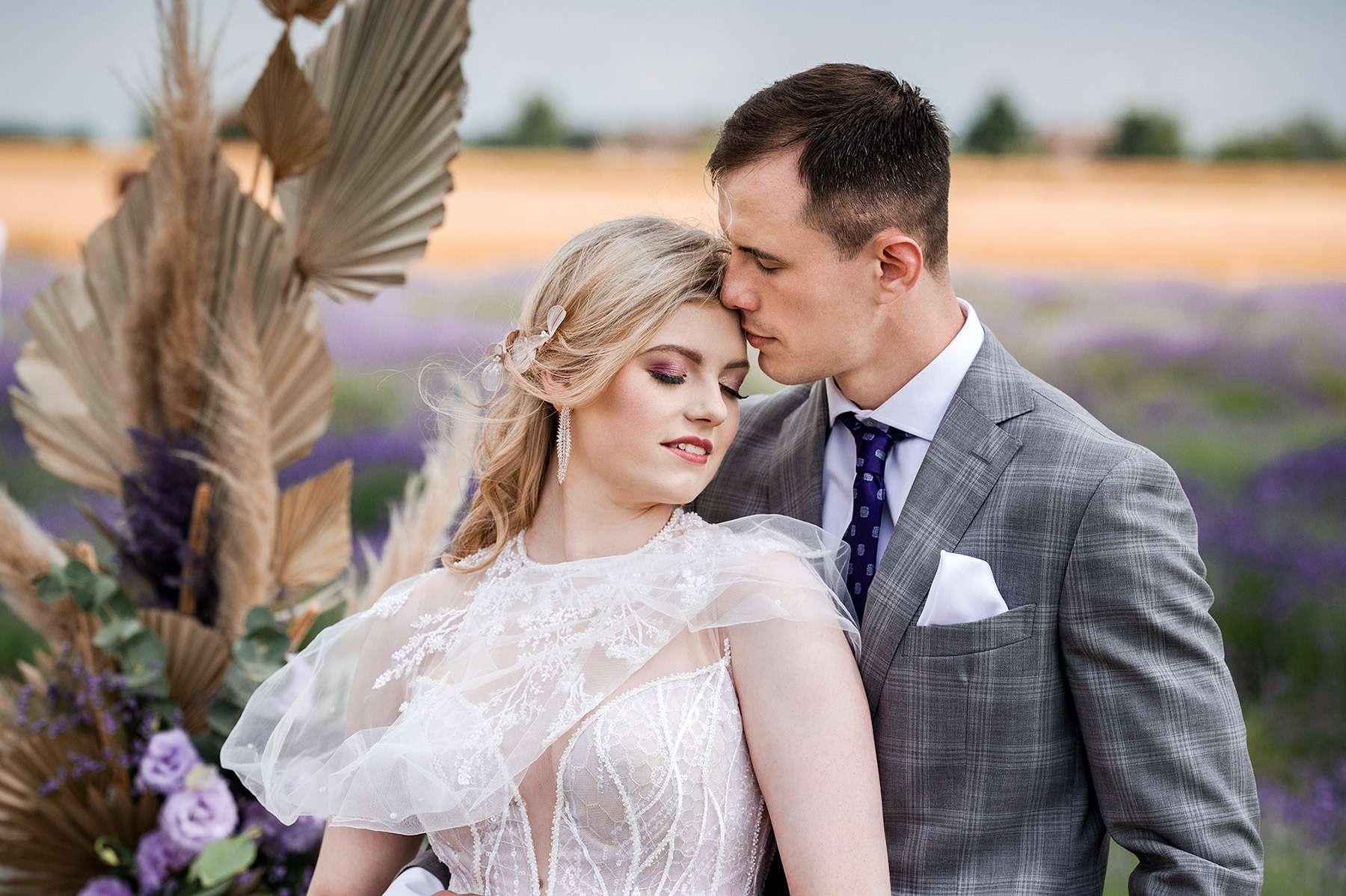 Para młoda w lawendowym polu: Panna młoda w koronkowej sukni z delikatnym makijażem, pan młody w szarym garniturze, czule dotykający czołem głowy ukochanej. Dekoracja z suszonych traw i fioletowych...