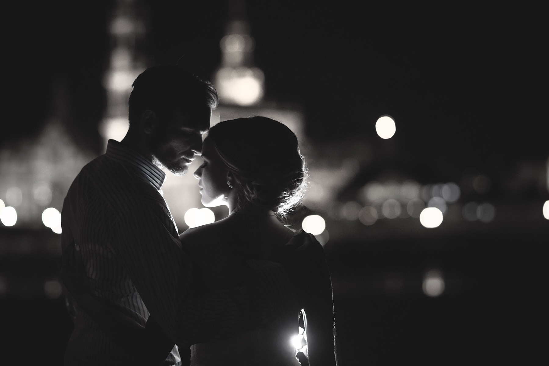 Czarno-białe zdjęcie pary w objęciach na tle nocnych świateł miasta, z silnym kontrastem i widocznymi sylwetkami.