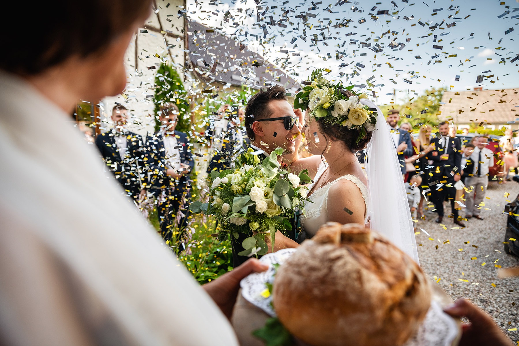 Para młoda w okularach słonecznych, obsypywana konfetti, z bochenkiem chleba na pierwszym planie, w tle goście weselni.