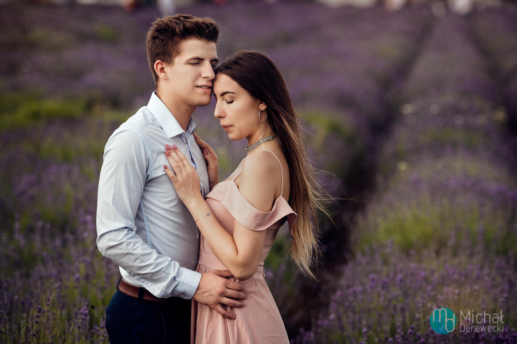 Zakochana para w objęciach na tle kwitnącej lawendy, on w błękitnej koszuli, ona w pudrowej sukience z chokerem i kolczykami kołami.
