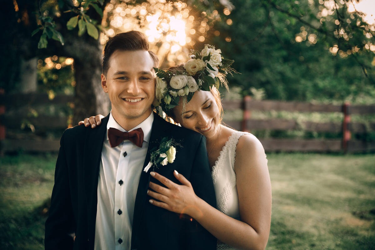 Uśmiechnięta para młoda w plenerze: pan młody w czarnym garniturze z bordową muchą, panna młoda w białej sukni z kwiatowym wiankiem opiera głowę na jego ramieniu, w tle drewniane ogrodzenie i zieleń.