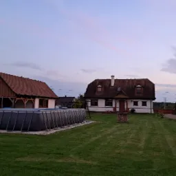 Ujęcie dwóch budynków z czerwonymi dachami, jeden z werandą, obok basen stelażowy i studnia, na tle wieczornego nieba.