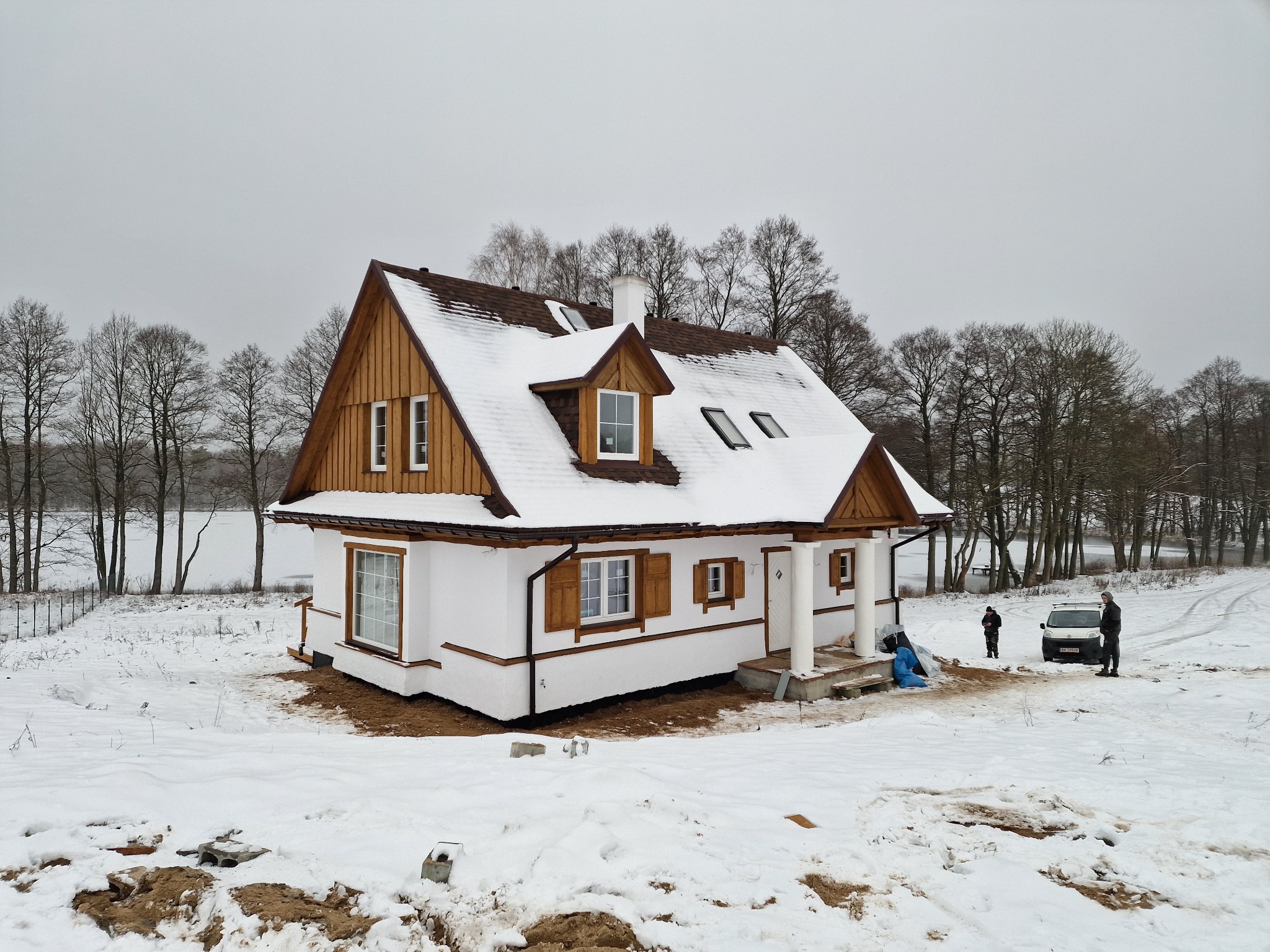 Zimowy krajobraz w Hajnówce: Nowo wybudowany dom z białymi ścianami i brązowymi oknami, częściowo pokryty śniegiem, z widocznym samochodem i dwiema osobami w tle.