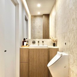 PROJEKTORNIA.COM.PL - Wąska łazienka z marmurowymi płytkami, wiszącą muszlą klozetową, umywalką na szafce z lamelami i podświetlanym lustrem.