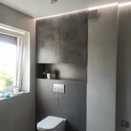 Nowoczesna łazienka z podwieszaną toaletą, szarą okładziną ścienną imitującą beton, oświetleniem LED podsufitowym i zabudową meblową z półką na roślinę doniczkową.