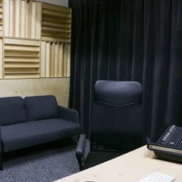 Wnętrze pomieszczenia z panelami akustycznymi na ścianie, ciemną sofą, krzesłem biurowym oraz sprzętem audio na biurku, zasłonięte czarnymi zasłonami.