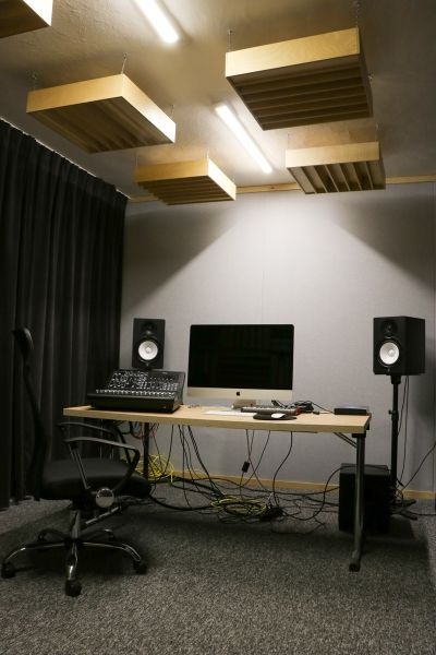 Wnętrze studia nagrań z biurkiem, komputerem, mikserem, głośnikami i drewnianymi panelami akustycznymi na suficie, widoczne kable pod biurkiem.