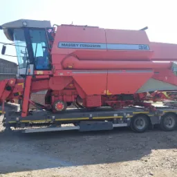 Czerwony kombajn zbożowy Massey Ferguson 32 przewożony na niskopodwoziowej lawecie, widok z boku na szarej, żwirowej powierzchni.